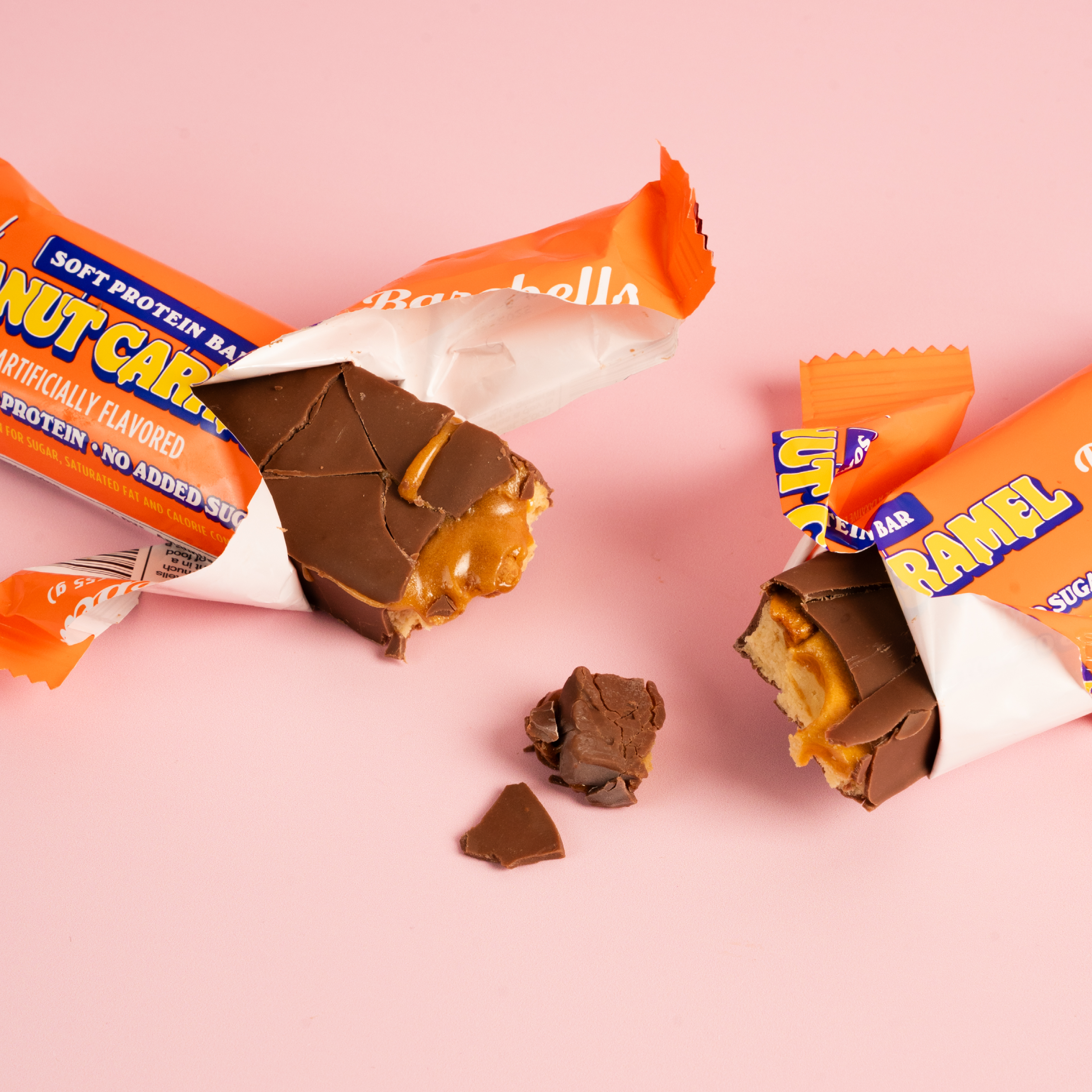 Protein bars with one partially unwrapped, revealing caramel and nuts on a pink surface.